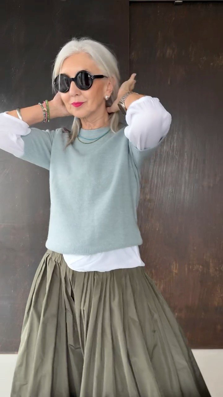 Silver-haired woman posing in a light blue sweater, white shirt, and olive green pleated skirt.