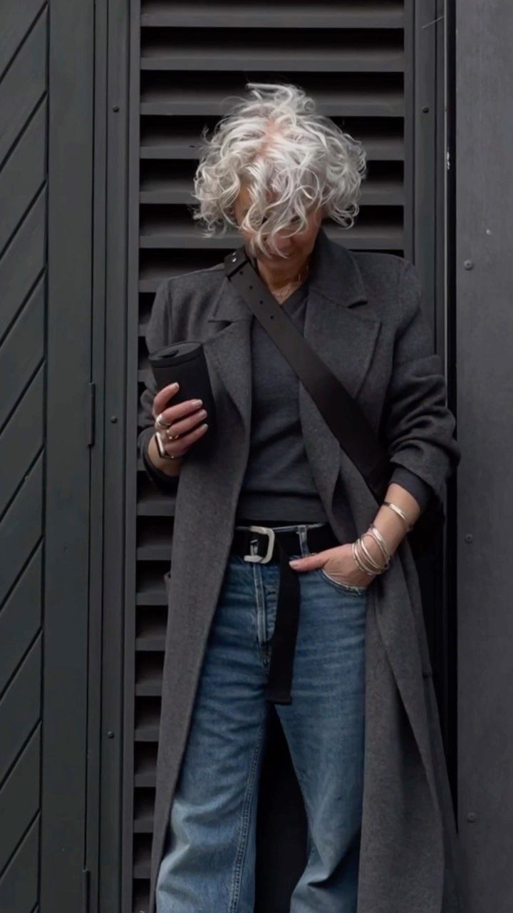 A woman with curly white hair wearing a long grey coat and blue jeans.