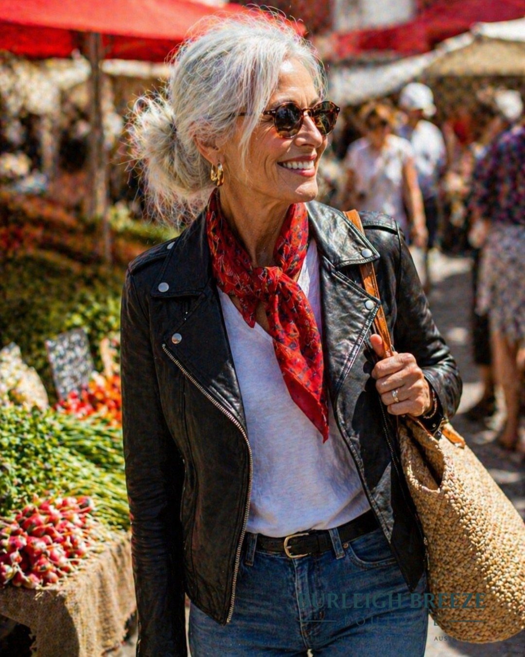 A smiling woman with grey hair wearing a black leather jacket and red scarf at a market.