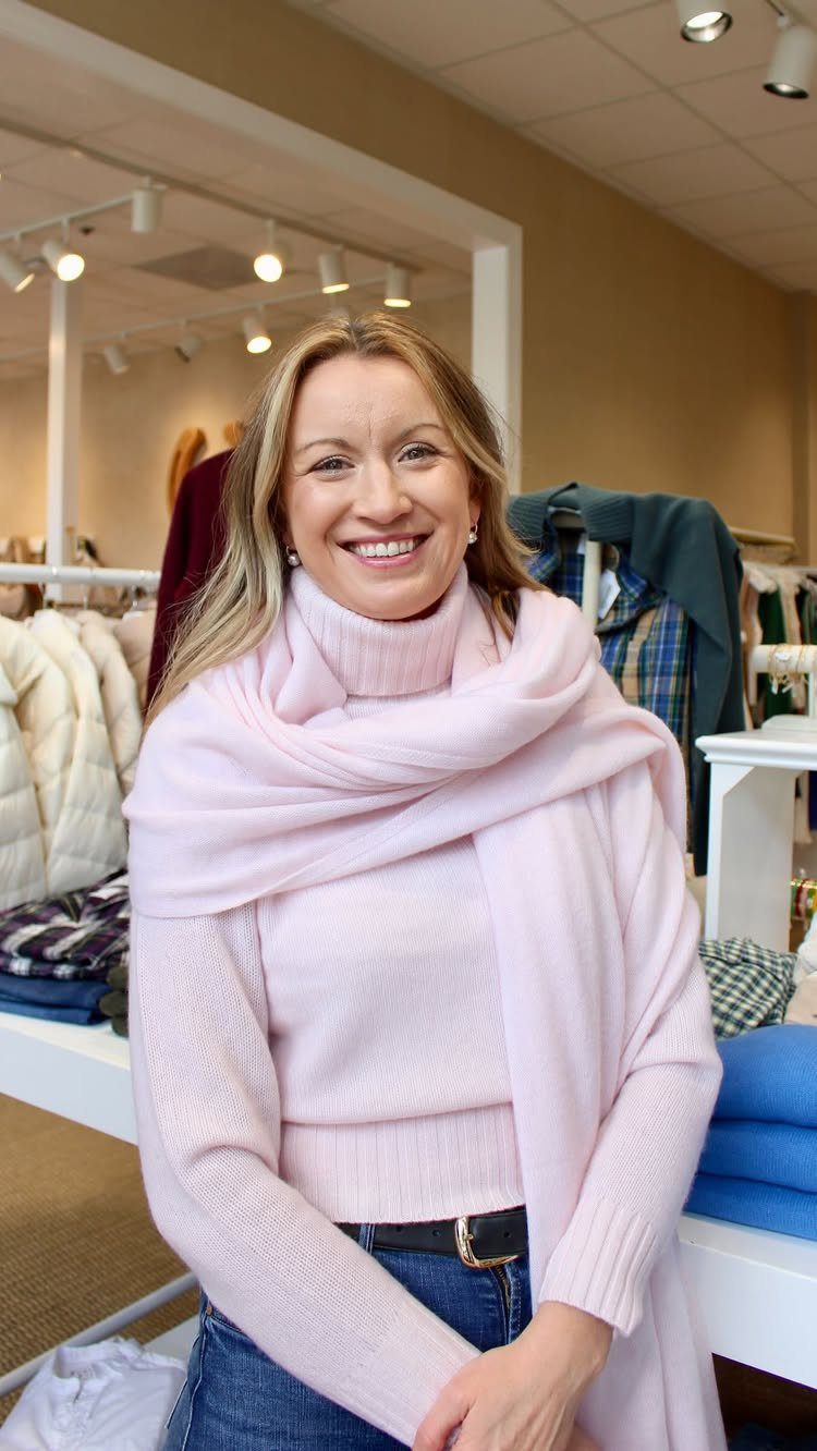 A smiling blonde woman wearing a pink turtleneck sweater and a matching pink scarf.