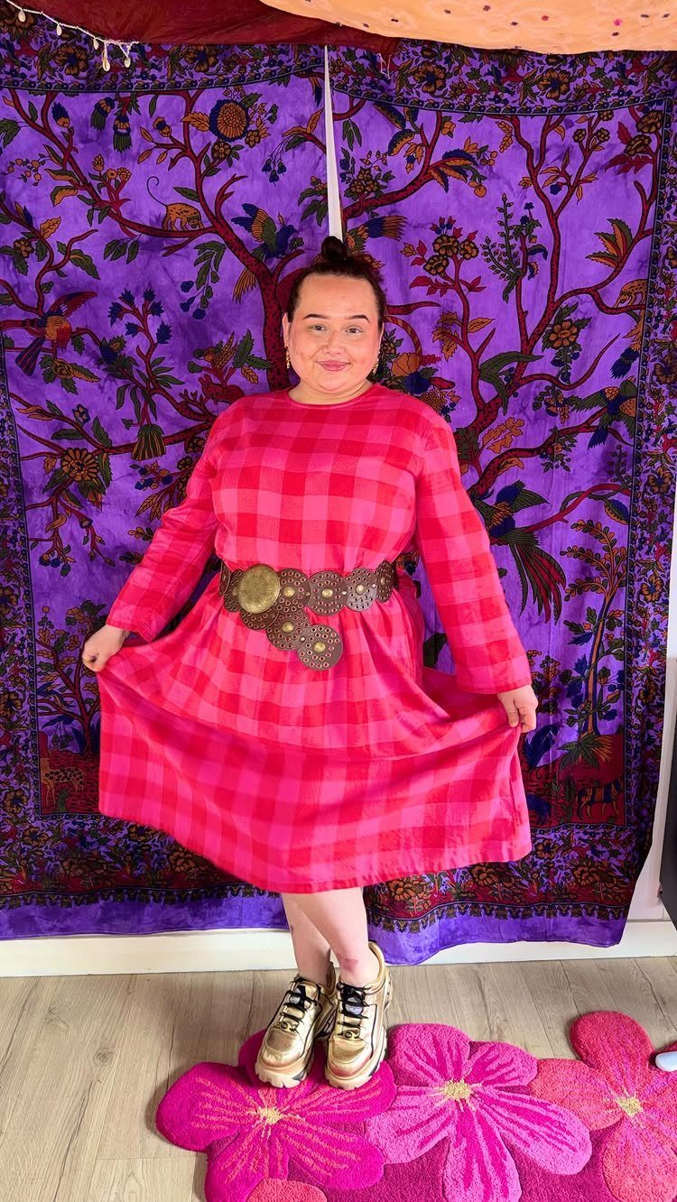 A woman wearing a pink checkered midi dress, a wide belt, and gold trainers.