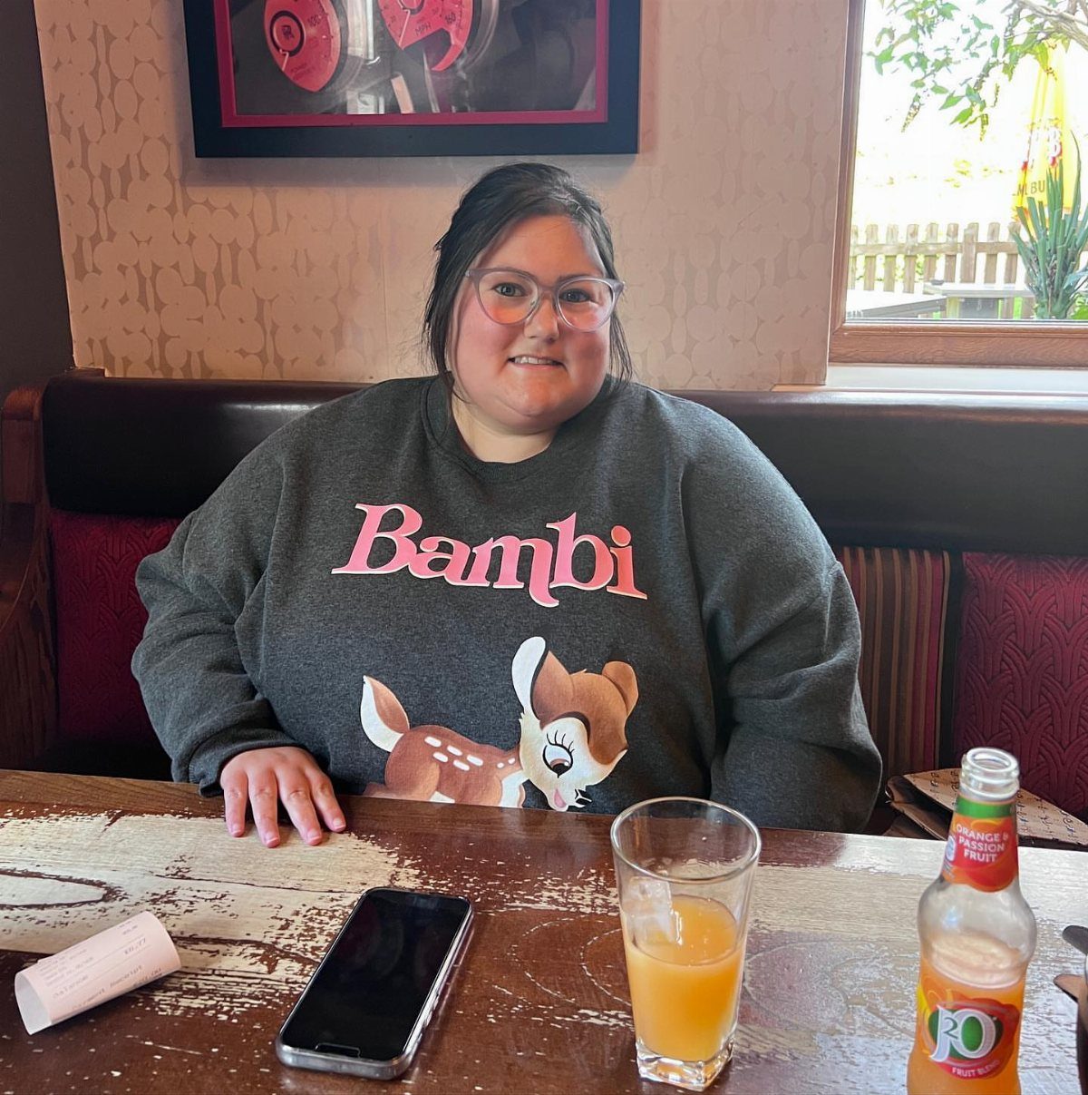 A smiling woman wearing a grey Bambi graphic crew neck sweatshirt sitting at a restaurant table.