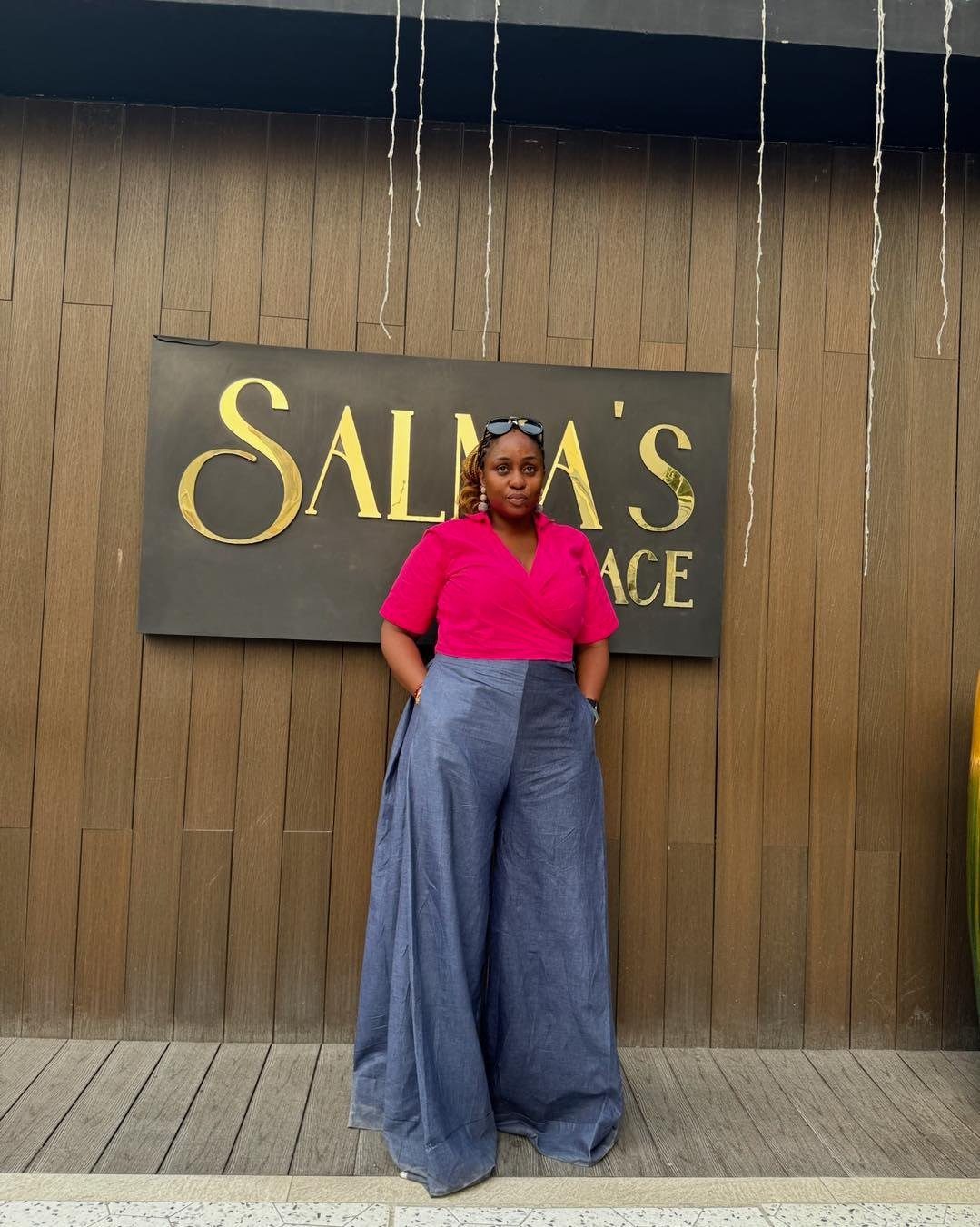 A woman wearing a pink fitted top and blue wide-leg trousers standing outdoors.