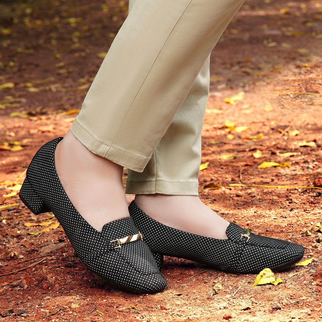 Polka dot black loafers with gold accents worn with beige trousers on a leaf-strewn ground.