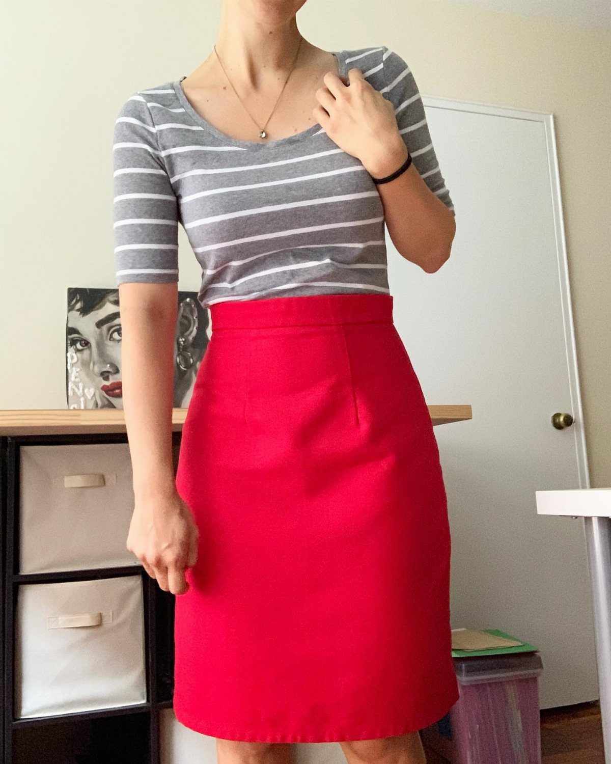 A person wearing a grey striped shirt and a vibrant red midi skirt standing in a room.
