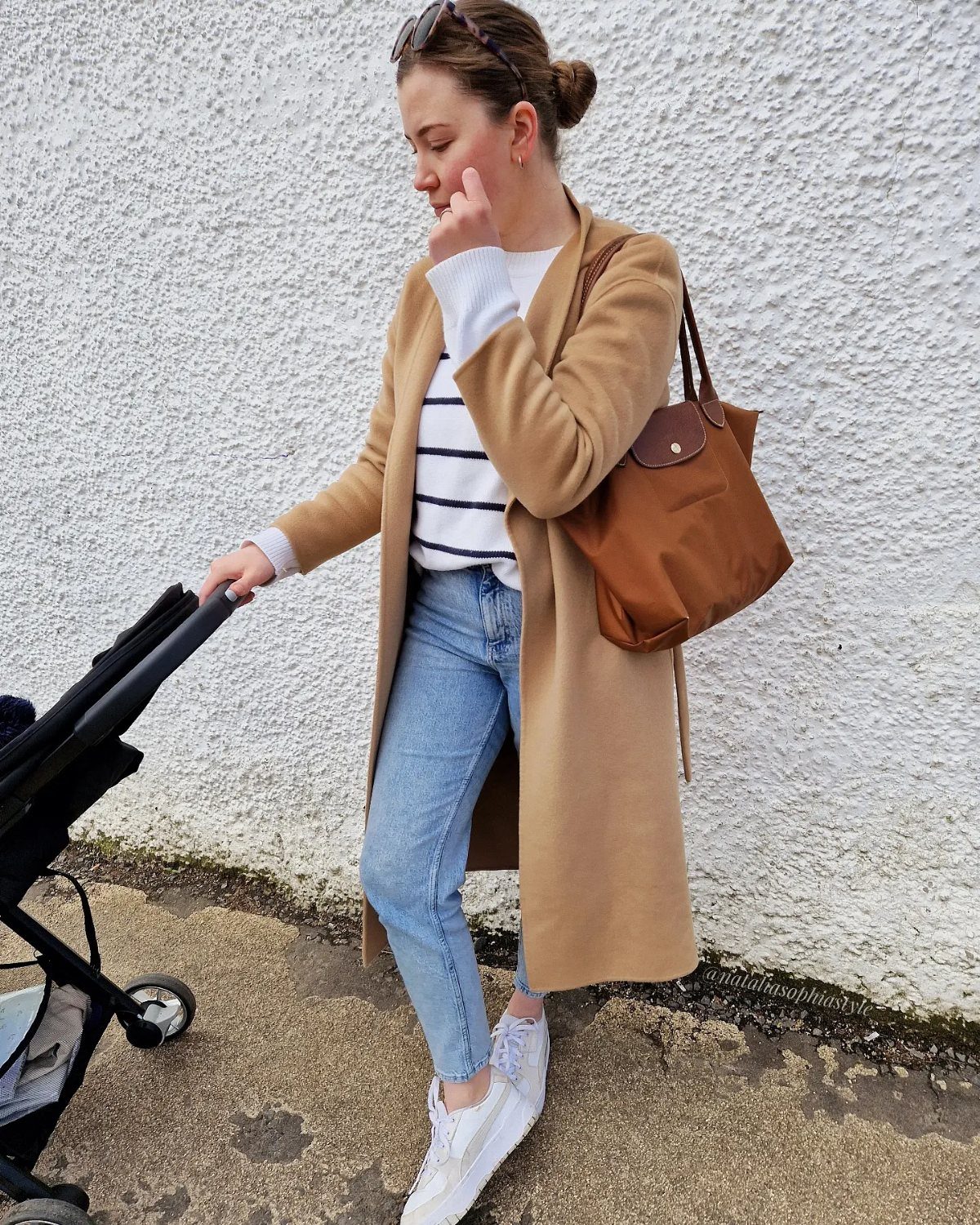 A woman in a tan coat, striped sweater, and jeans walking with a stroller.