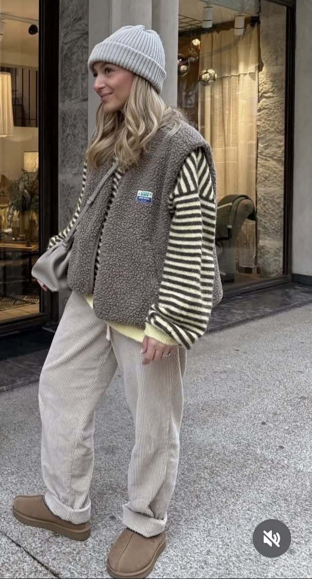 A woman in a striped shirt, fleece vest, and corduroy trousers standing on a city street.