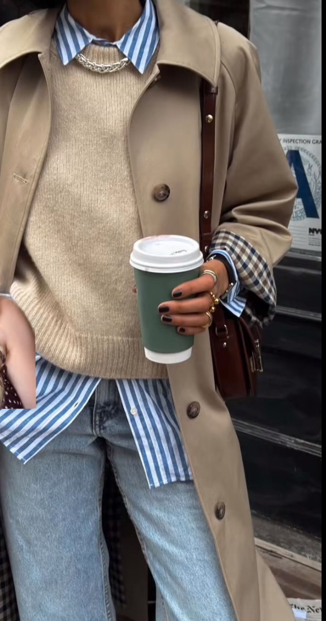 A person wearing a beige trench coat, cream sweater, striped shirt, and blue jeans while holding coffee.
