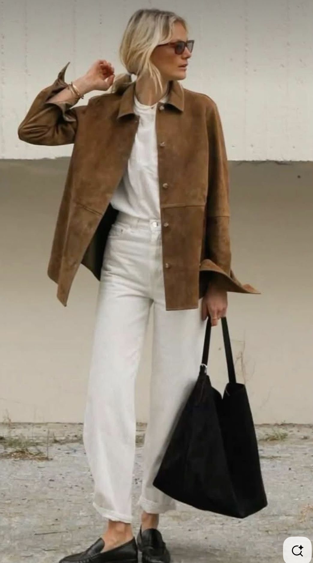 Blonde woman in white trousers and a brown suede jacket holding a black tote bag.