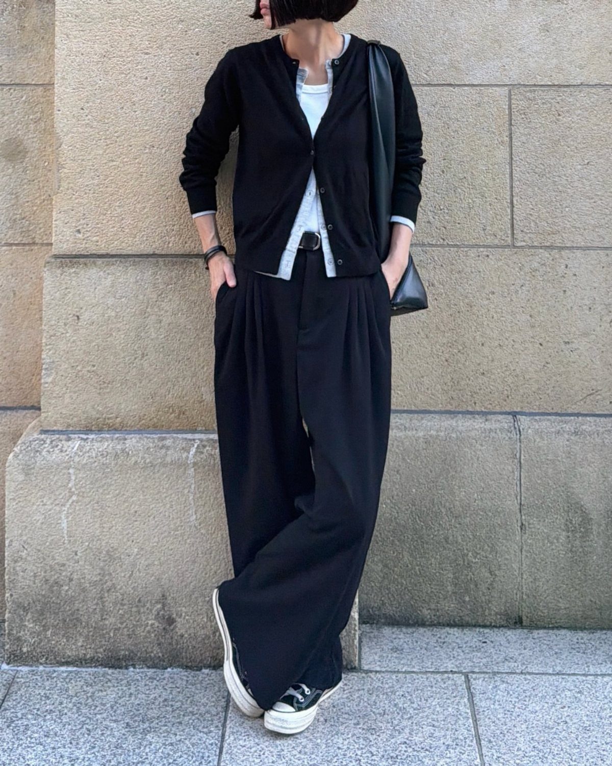 Person wearing a black cardigan, white top, wide-leg black trousers, and black sneakers against a stone wall.