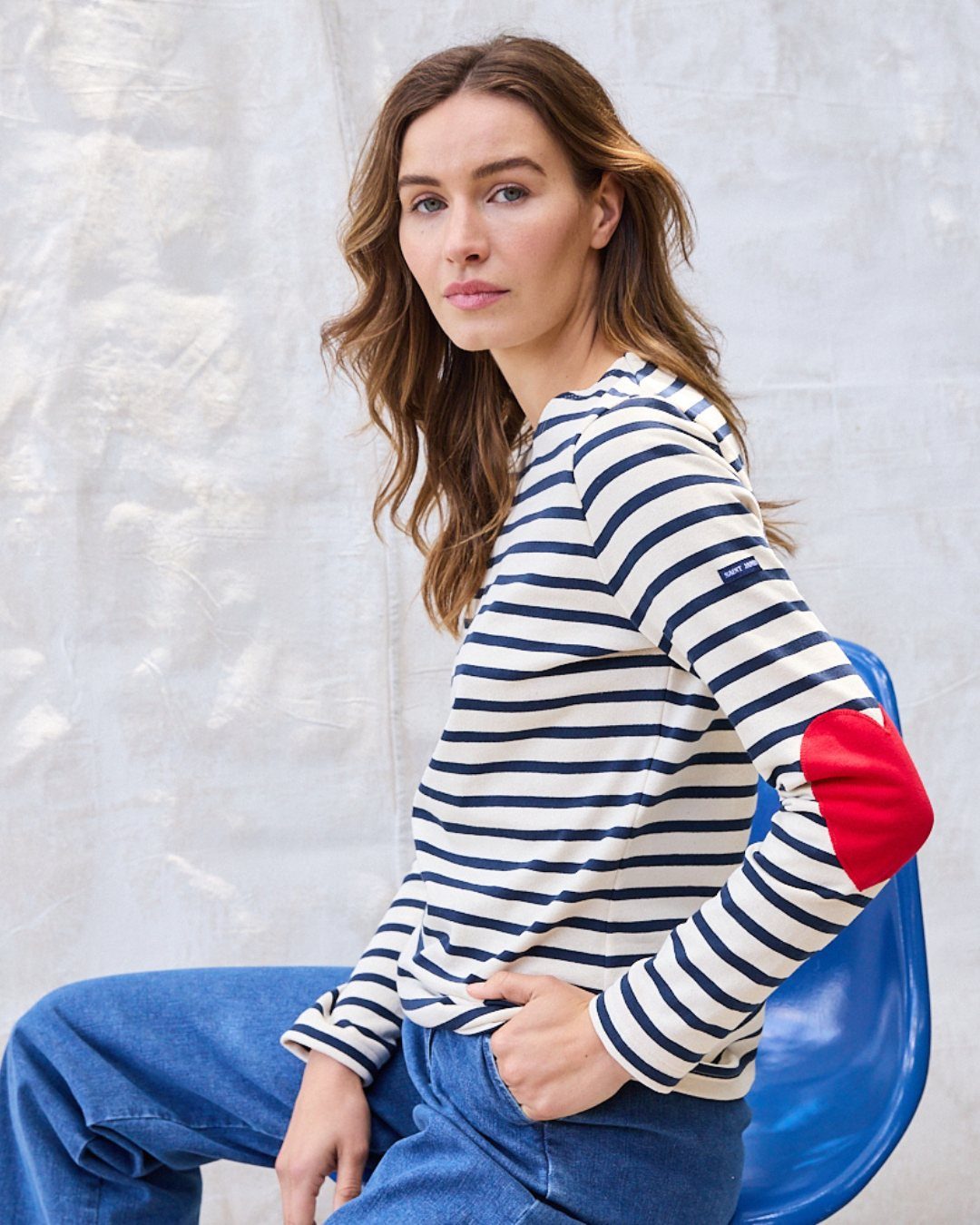 A woman wearing a navy and white striped Breton shirt and blue jeans sitting on a blue chair.