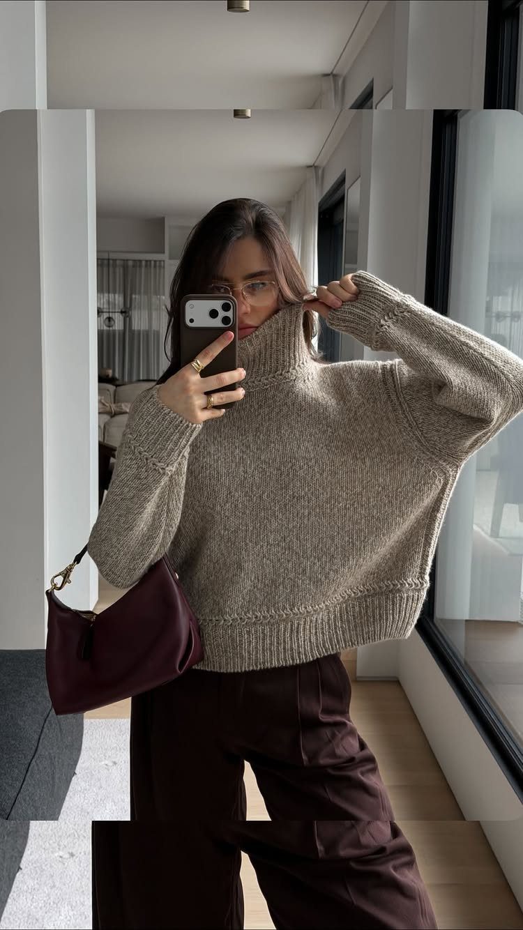 A woman posing in a mirror wearing a thick beige knit sweater and burgundy trousers.