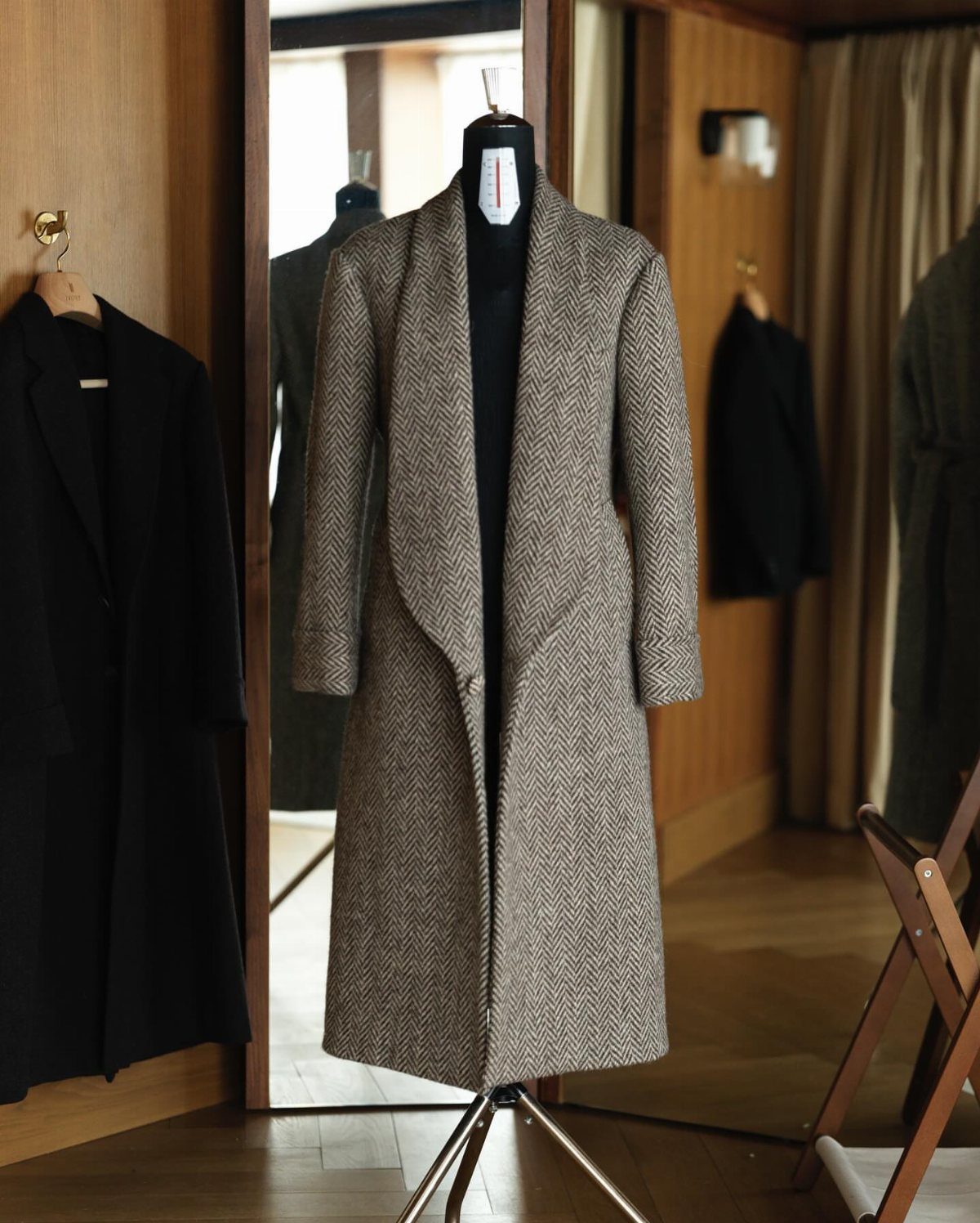 A long grey herringbone wool coat displayed on a black mannequin in a wooden room.