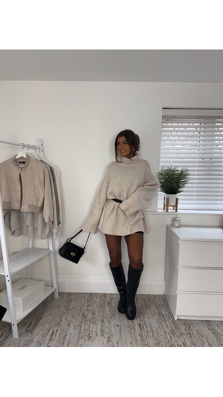 Woman wearing a beige oversized sweater, black shorts, and black knee-high boots standing in a room.