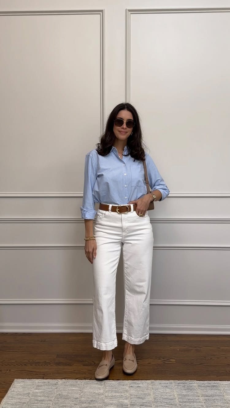 A woman wearing a blue shirt, white trousers, and beige loafers standing against a wall.
