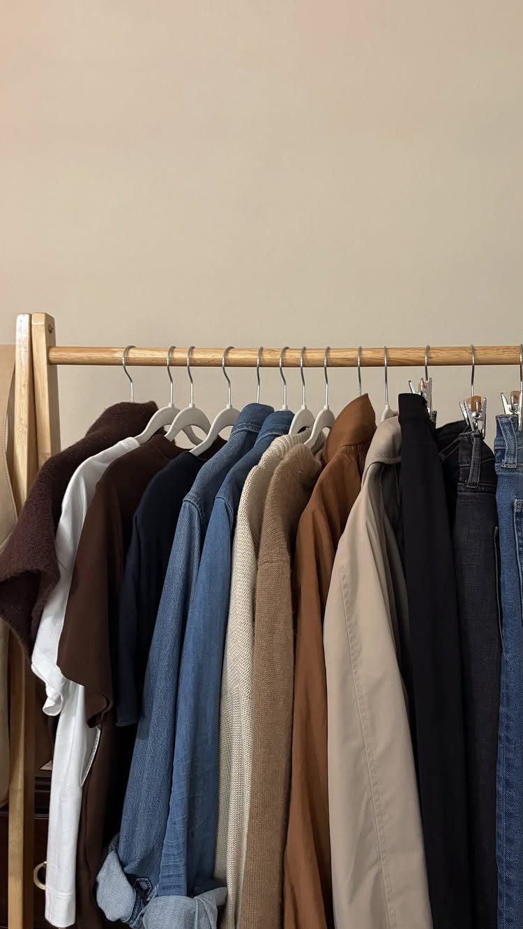 A wooden clothing rack displays a collection of neutral-toned shirts and jackets on white hangers.