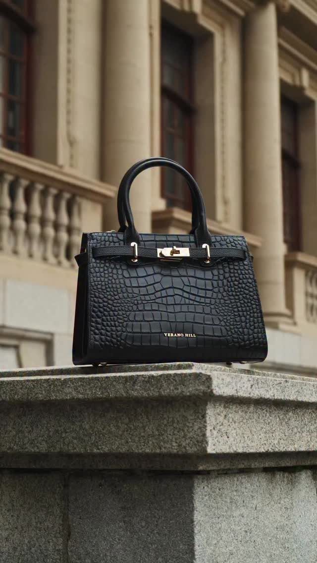 Black crocodile-textured structured handbag with gold hardware resting on a stone ledge outdoors.