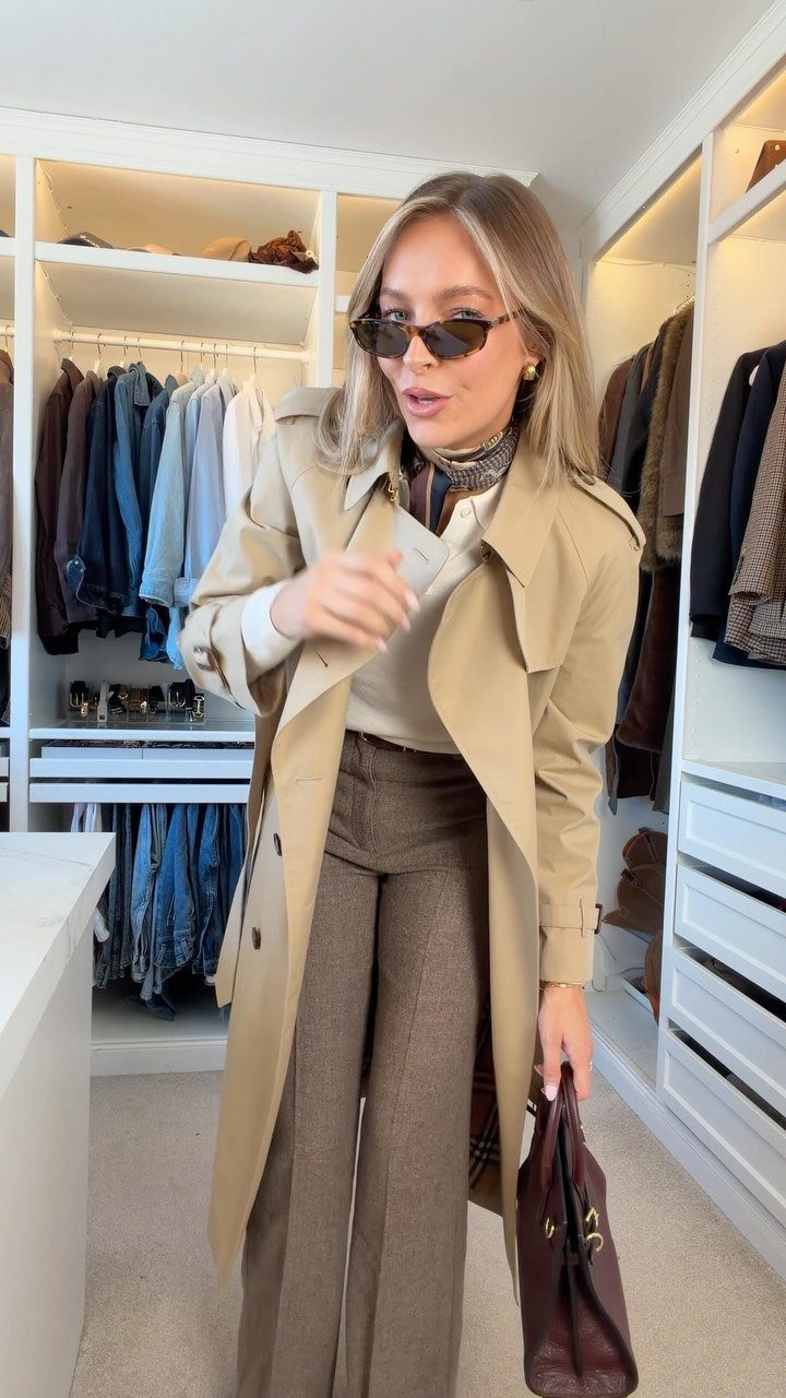 Woman wearing a beige trench coat, cream sweater, and brown trousers in a closet.