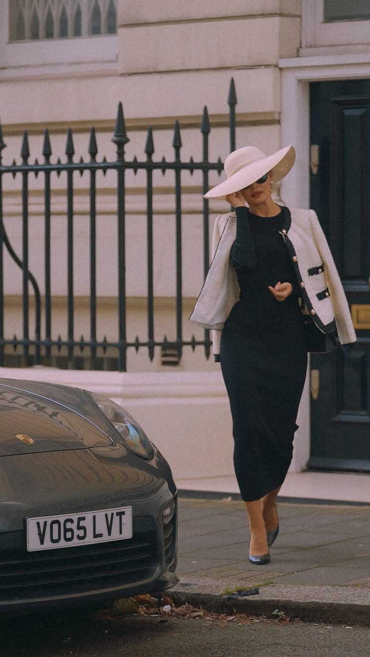 Woman in a long black dress, white hat, and draped blazer walking on a city street.