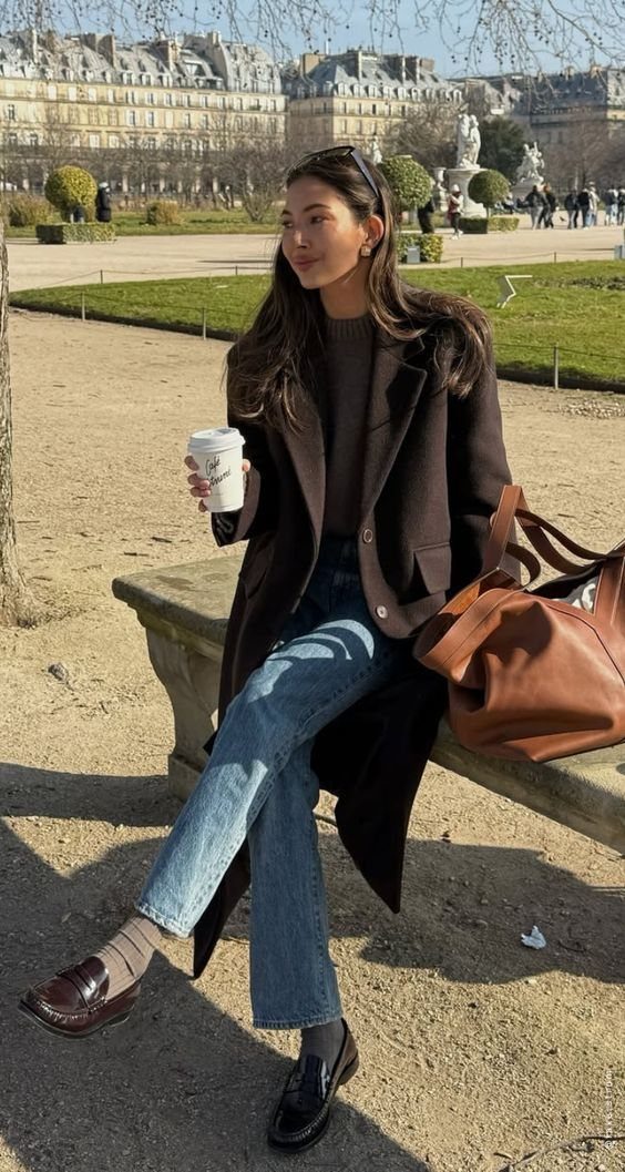 A woman sitting on a park bench wearing a long brown coat, blue jeans, and black loafers.