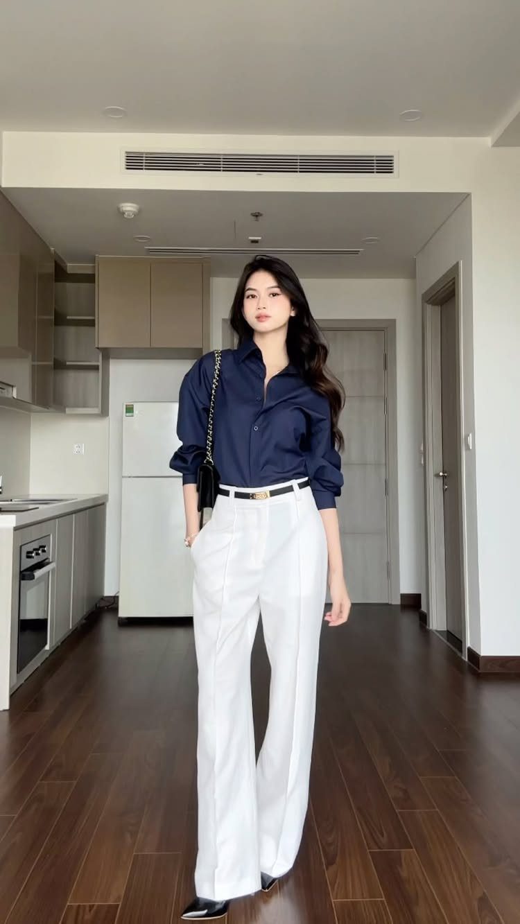 Woman posing in a navy button-down shirt and white wide-leg trousers in a modern apartment.