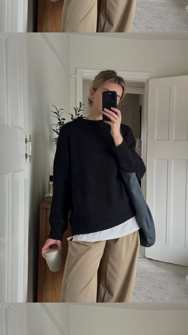 Woman in a black knit sweater and beige straight trousers taking a mirror selfie.
