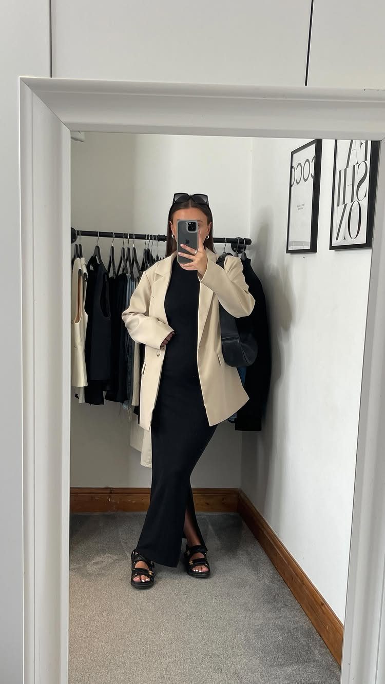 Woman in a black maxi dress and cream blazer posing for a mirror selfie.