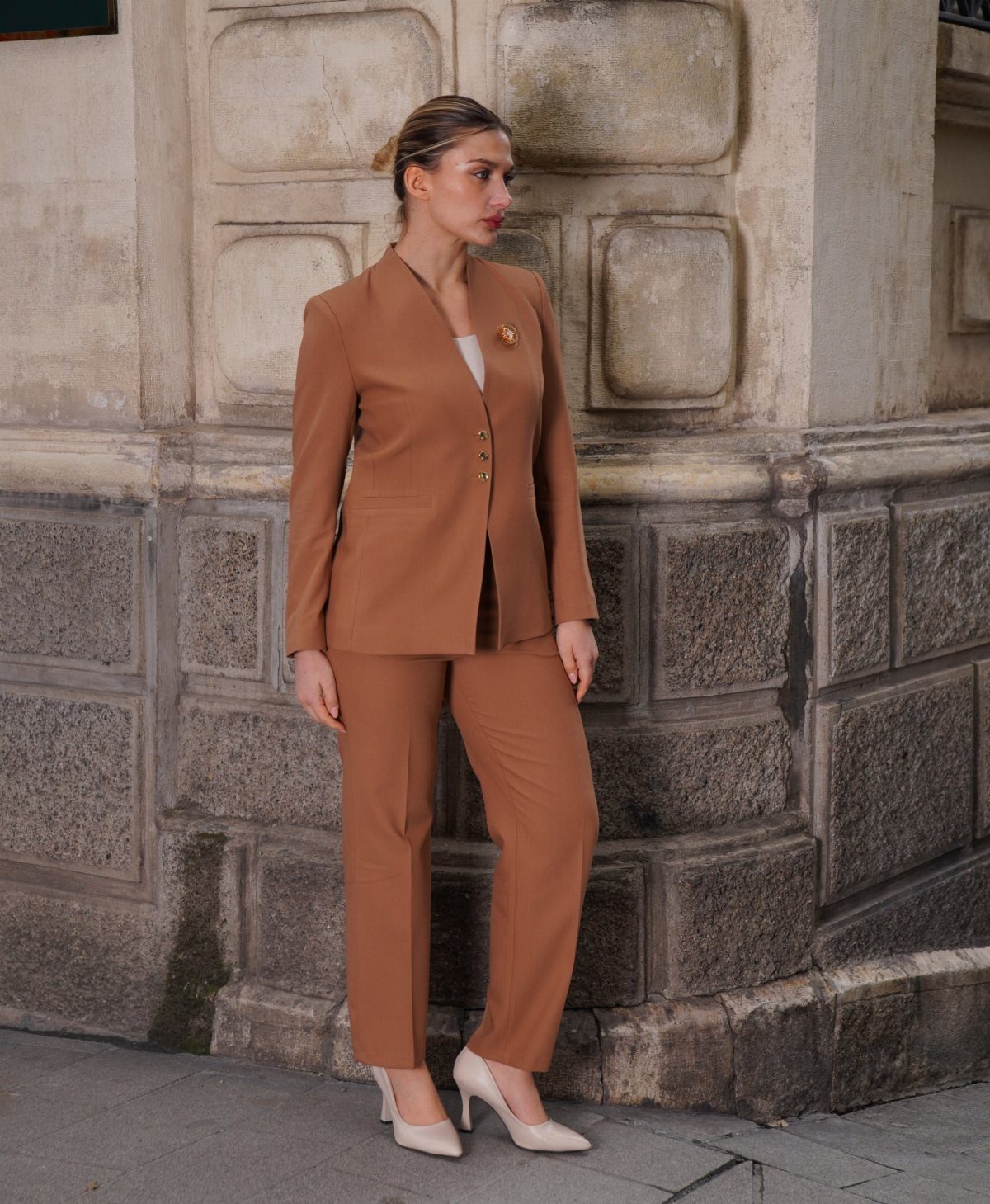 A woman wearing a terracotta tailored suit and cream heels standing against a stone wall.