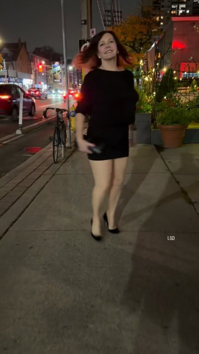 A woman in a short black dress and heels walking on a city sidewalk at night.
