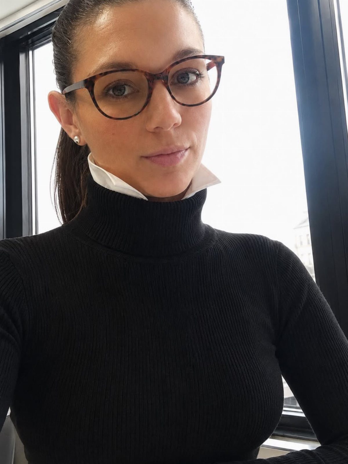 A woman wearing a black ribbed turtleneck sweater and tortoiseshell glasses indoors.