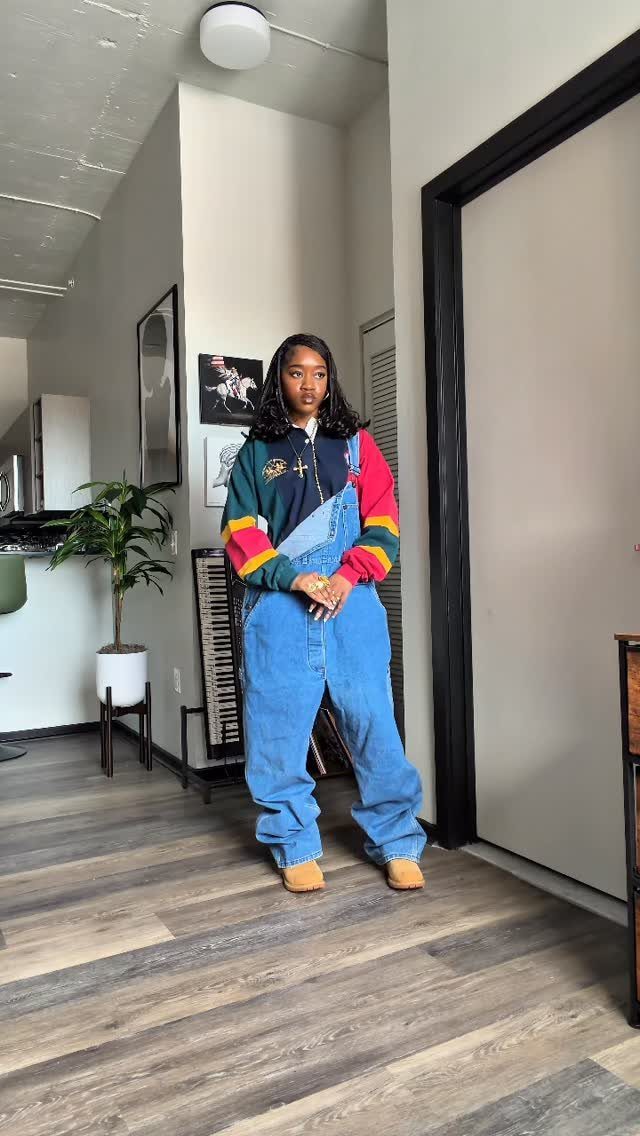 Woman posing in baggy denim overalls and a colorful vintage striped sweater indoors.