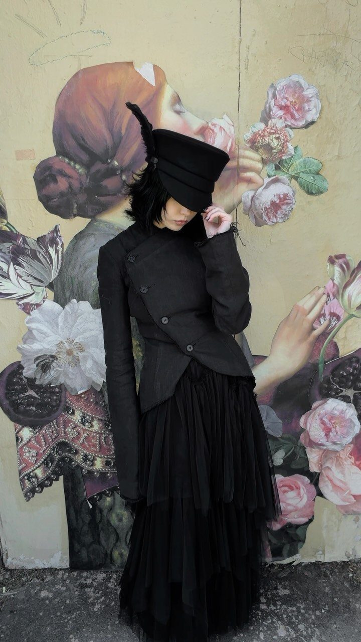 Woman in an all-black structured jacket and tiered tulle skirt posing before a floral mural.