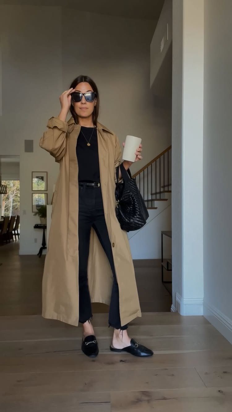 A woman in a tan trench coat, black top, and black trousers posing indoors.