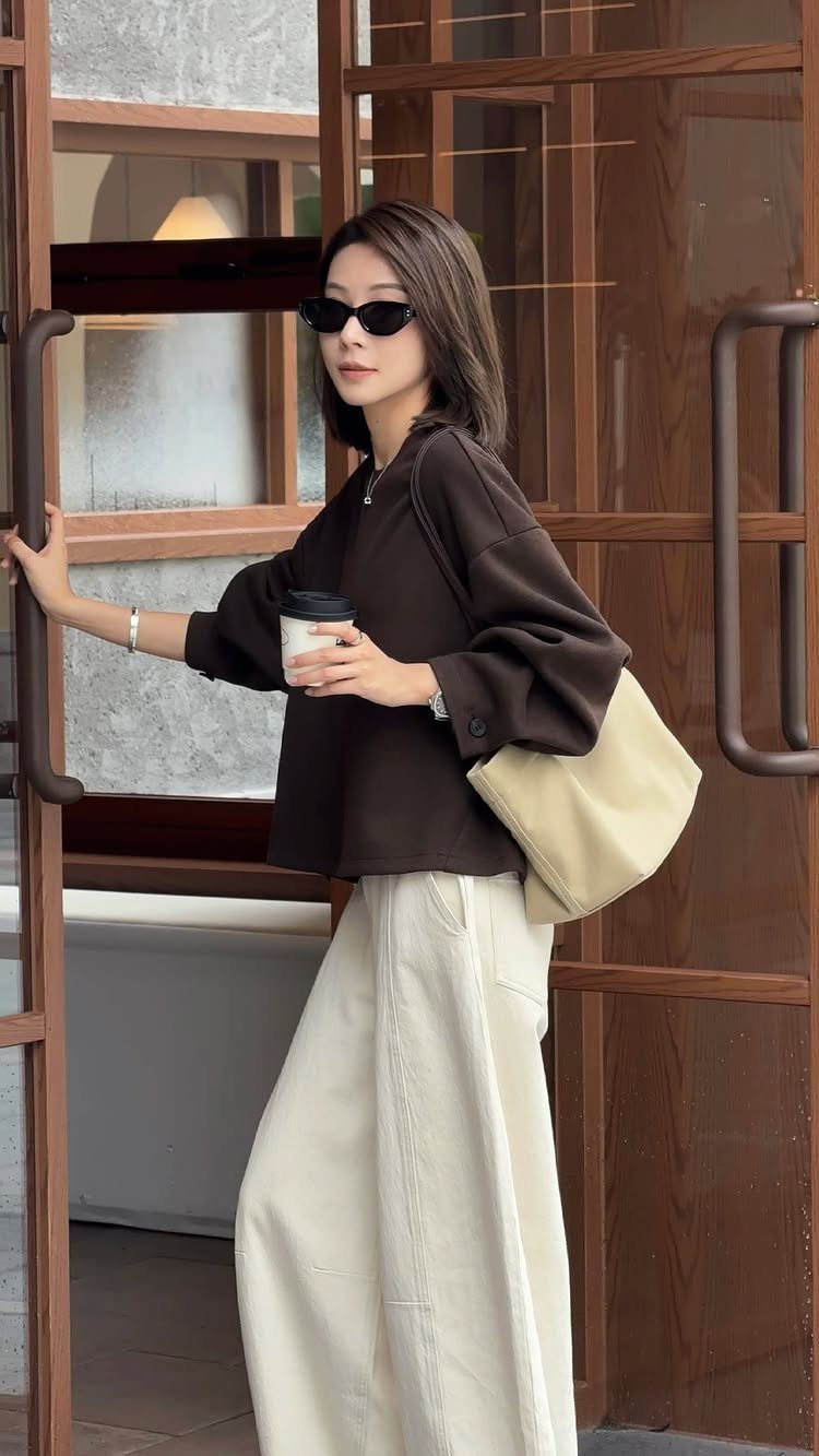 Woman in dark brown blouse and cream wide-leg trousers standing by a wooden door.