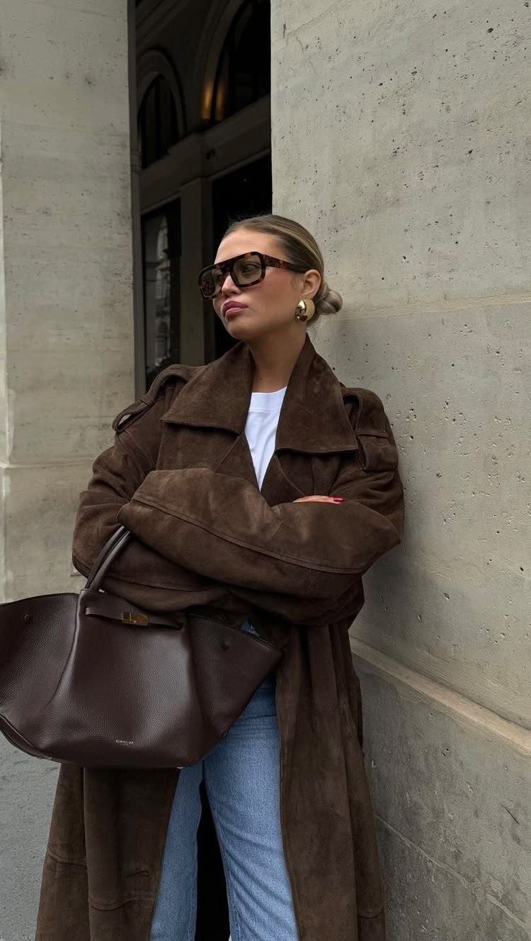 A woman in a long brown suede coat and blue jeans holding a large leather handbag.