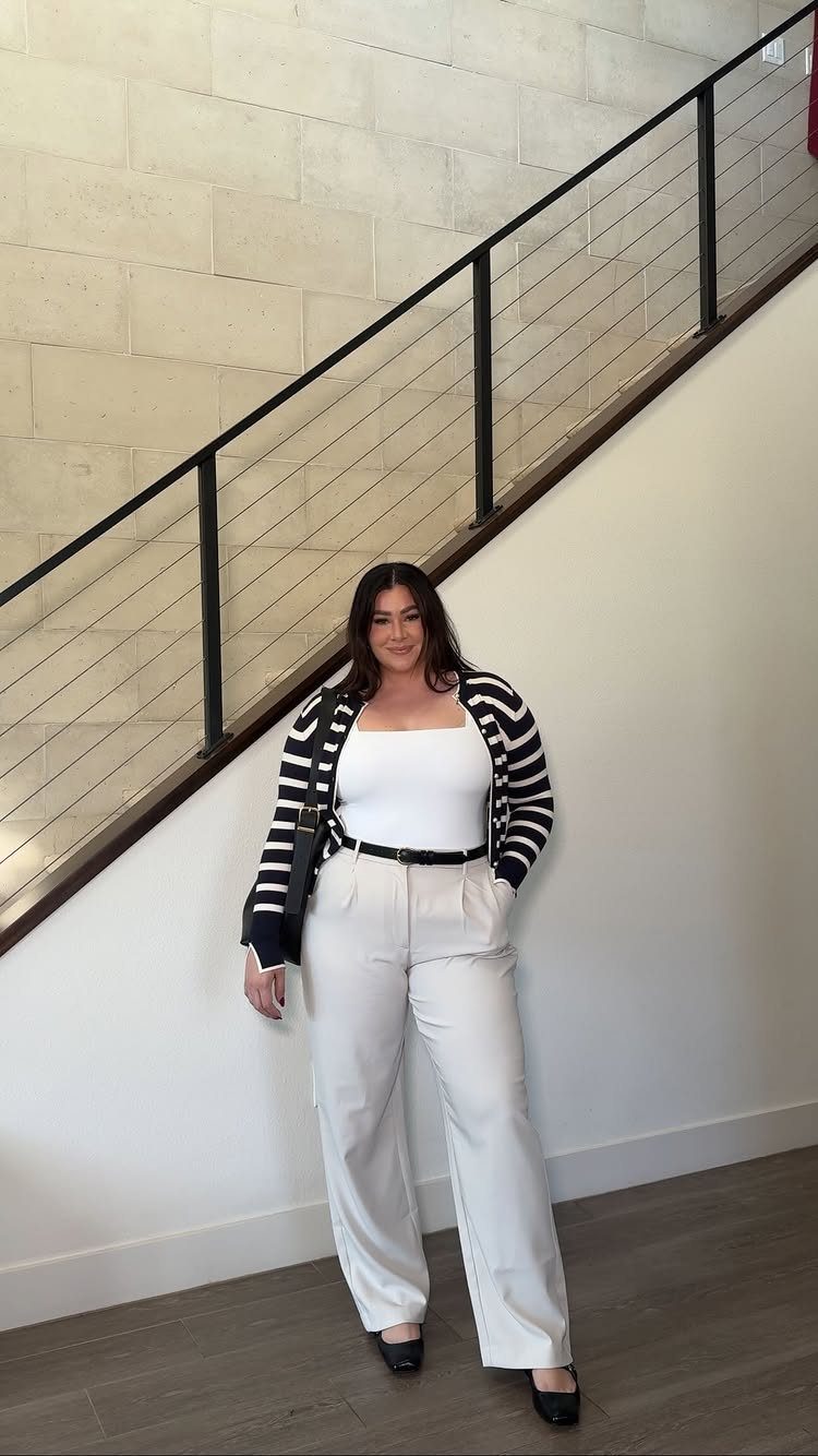 A plus-size woman posing in a striped cardigan and cream wide-leg trousers by a staircase.