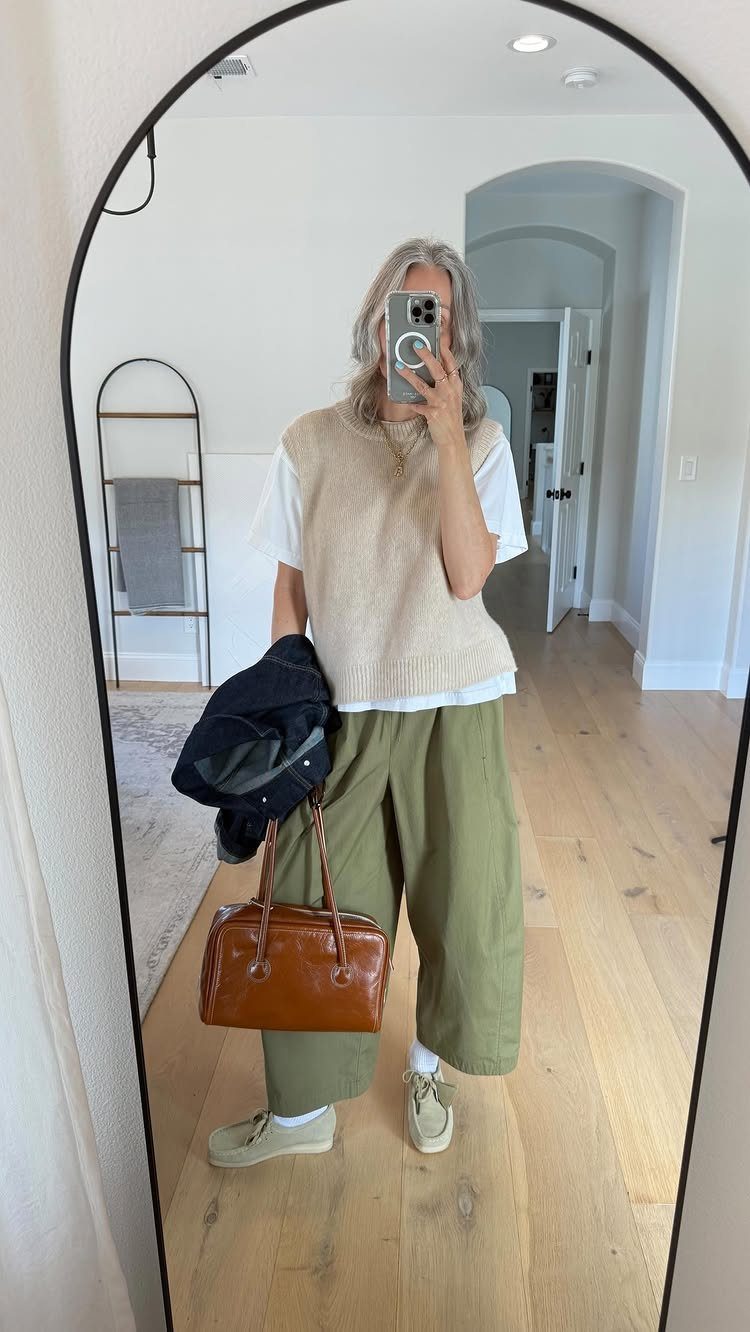 A person posing in a mirror wearing a beige sweater vest and olive green trousers.