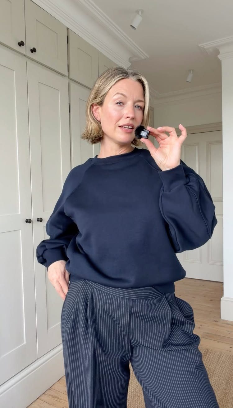 A blonde woman wearing a navy sweatshirt and pinstriped trousers holding a small cosmetic jar.