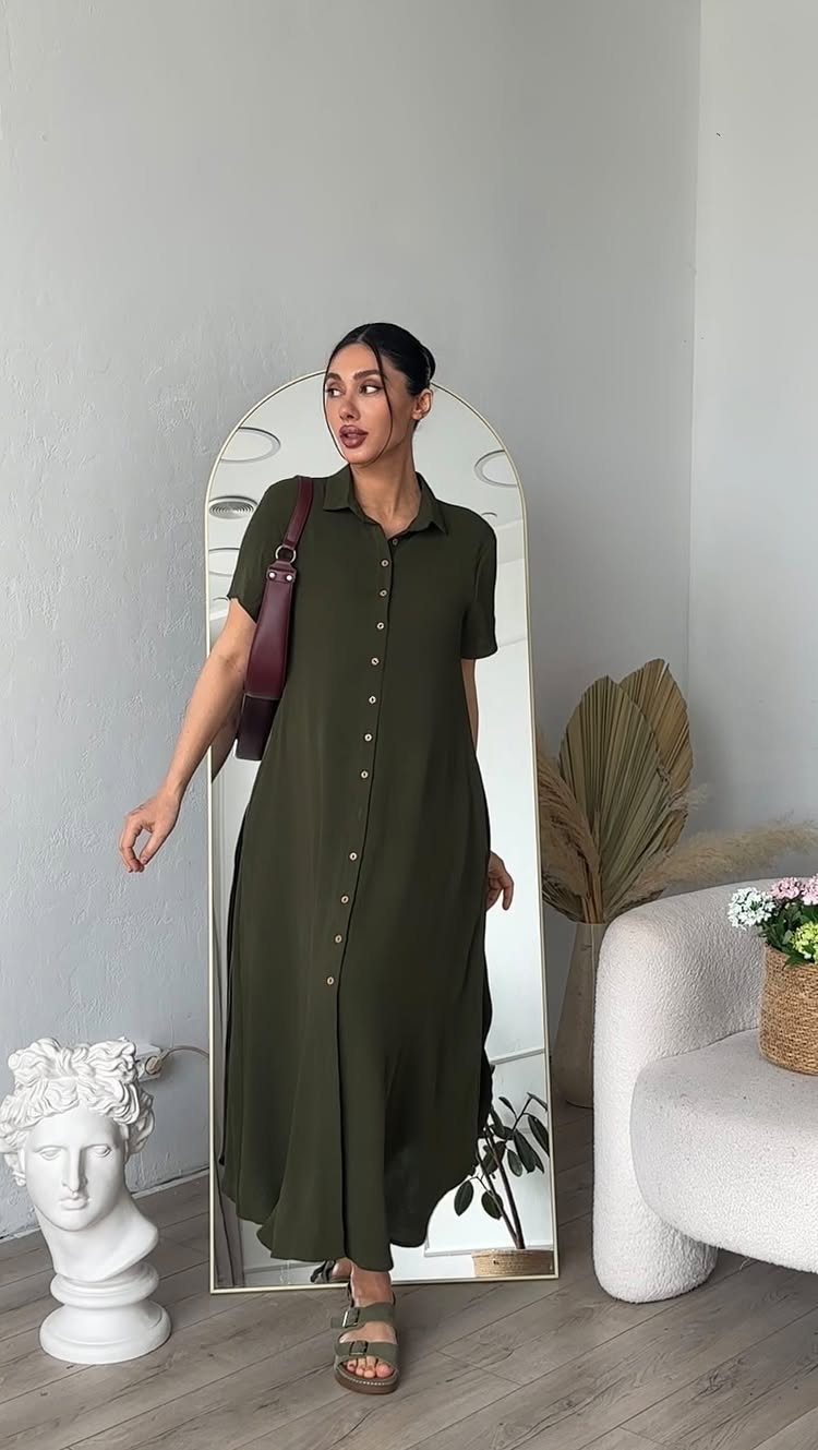 A woman wearing a long olive green button-down dress standing in a bright room.