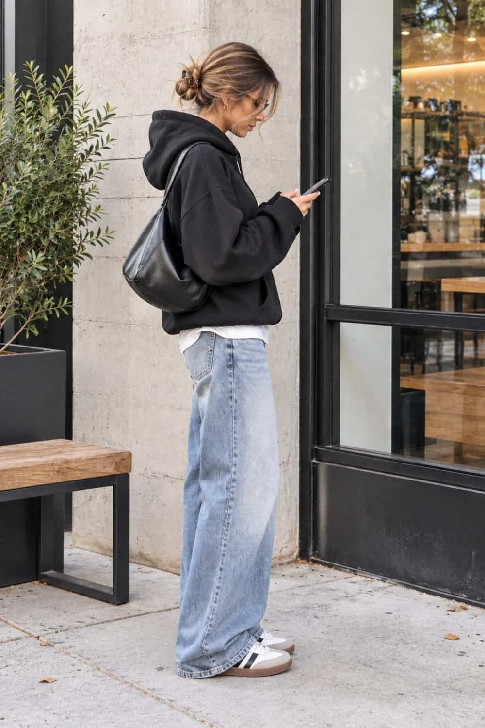 Woman in a black hoodie and wide-leg blue jeans standing outdoors using her phone.