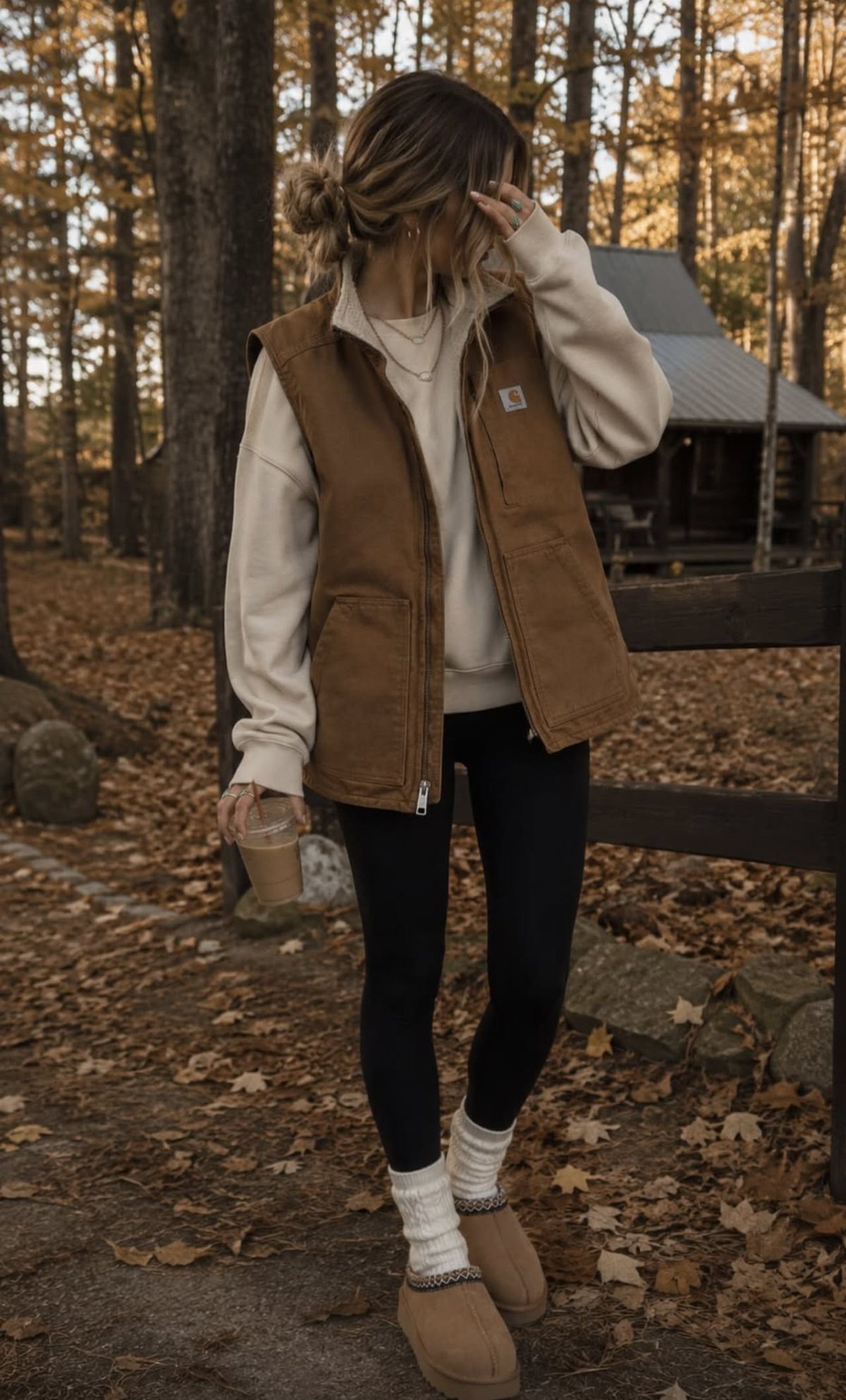 A person wearing a brown vest and cream sweatshirt standing in an autumn forest.