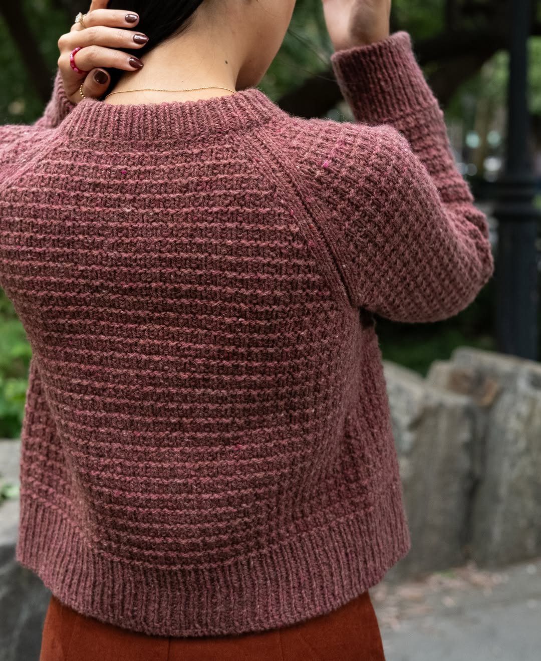 A person wearing a textured dusty rose cropped sweater and brown pants outdoors.