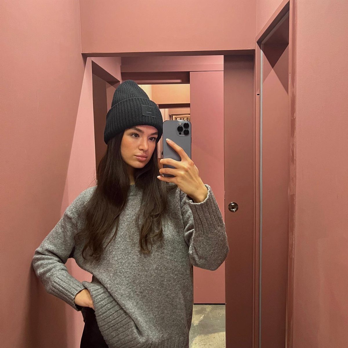 A woman in a grey oversized knit sweater and black beanie taking a mirror selfie.