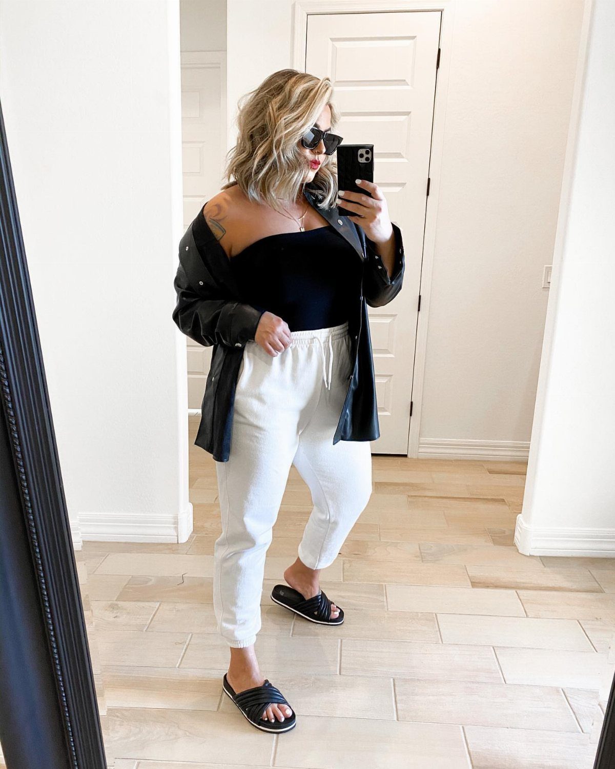 Woman in a black bodysuit, leather jacket, and white trousers posing for a mirror selfie.