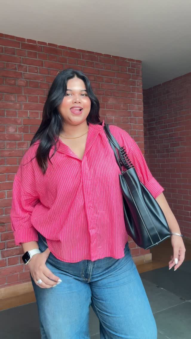 Woman in a pink striped button-down shirt and blue jeans standing by a brick wall.