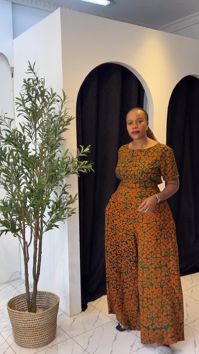 A woman posing in a patterned orange and green short-sleeved jumpsuit indoors.