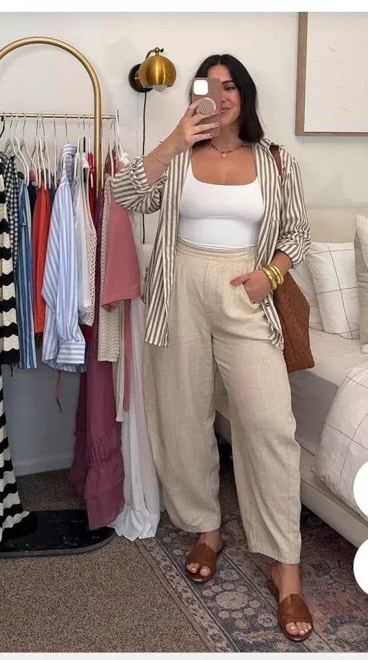 Woman wearing a white tank top, striped shirt, and beige linen trousers standing indoors.