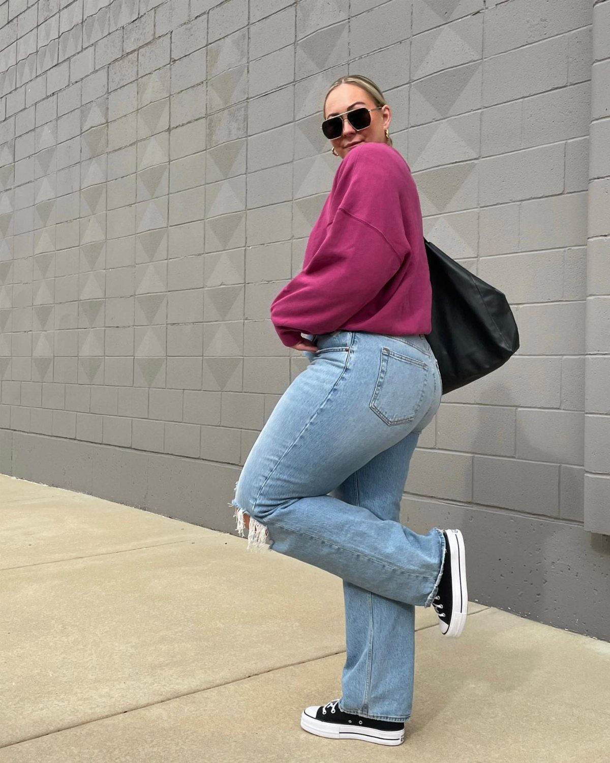Woman in a magenta oversized sweater and light blue jeans posing against a grey wall.