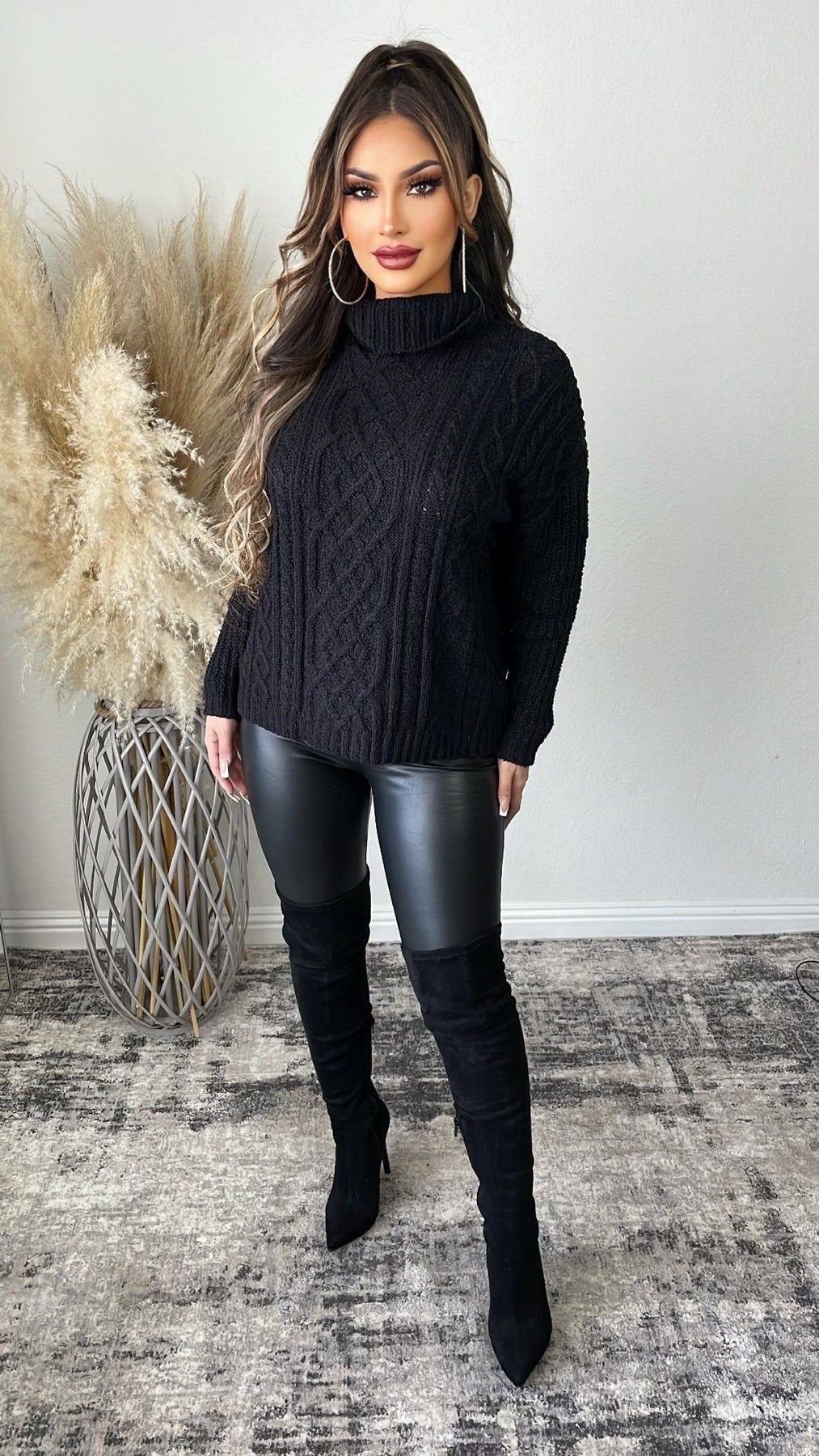 Woman posing in a black cable-knit turtleneck, black leggings, and tall black boots.
