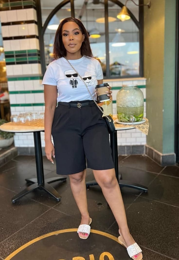 A woman wearing a white graphic t-shirt and black tailored shorts standing outdoors.